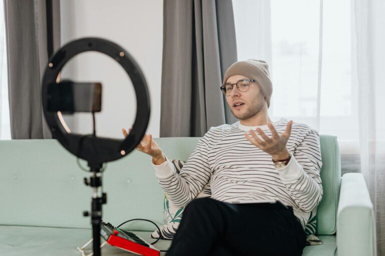 A man in a beanie using a ring light to vlog indoors on a couch.